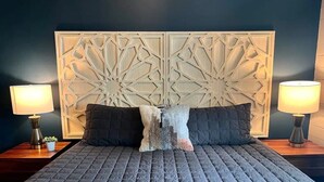 A bed with a quilted grey cover, two dark pillows, and a decorative headboard with geometric patterns. There are two bedside tables with lamps and a small plant on each. - Haus of Style Moody Blues (Charlotte)