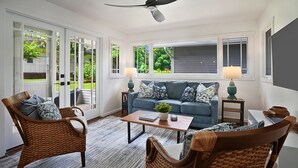 Un salón con muebles de mimbre, un ventilador de techo, un sofá con cojines estampados y vistas al verdor exterior.