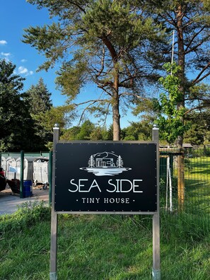 A sign for 'SEA SIDE Tiny House' with a logo of a house on water, surrounded by greenery and trees.