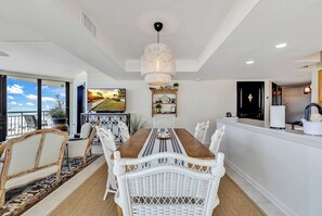 Un área de comedor con una mesa de madera grande, sillas de mimbre blanco, una lámpara colgante y un televisor en la pared.