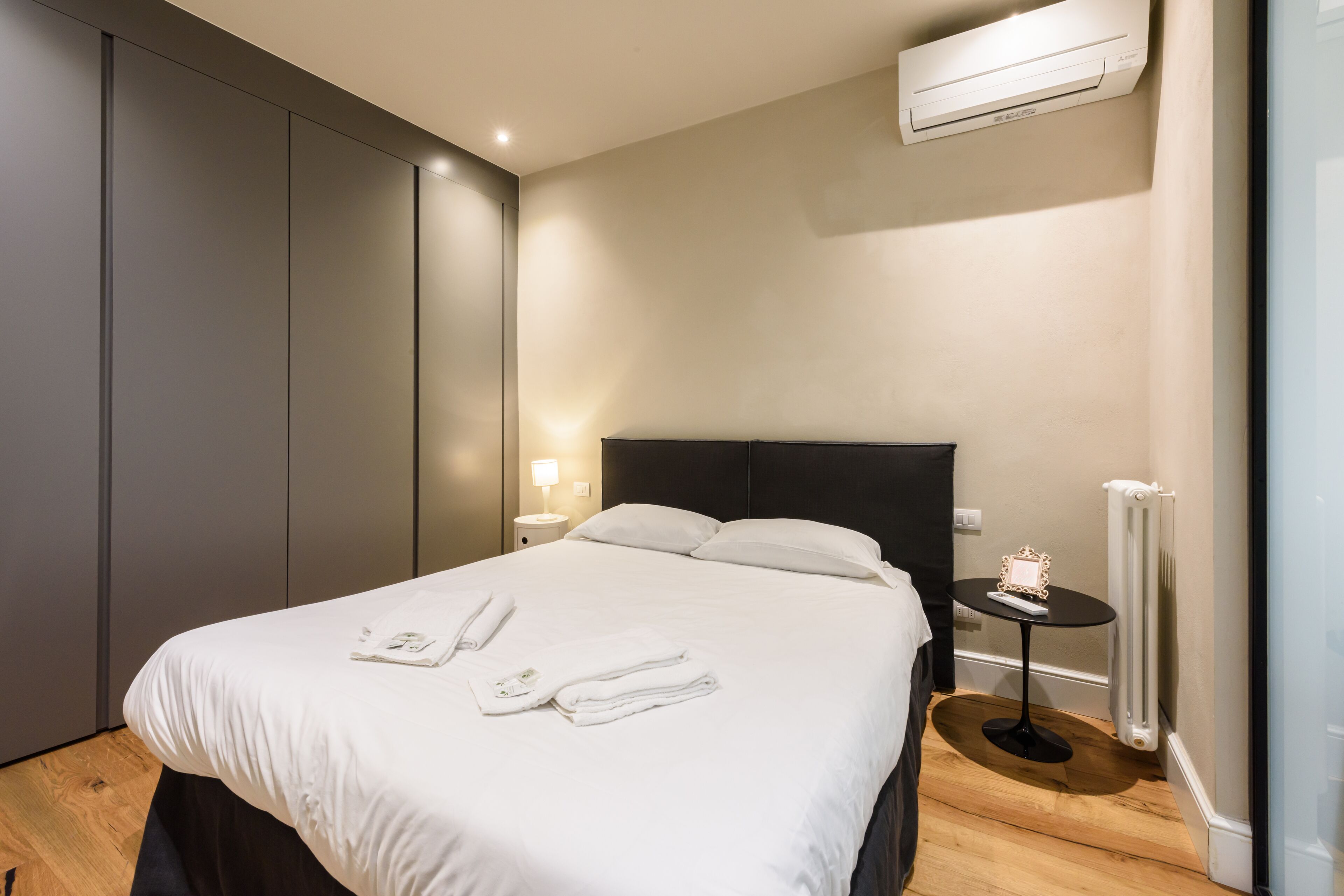 A hotel room with a large bed, a bedside table with a lamp, a small table with a picture frame, and an air conditioning unit on the wall.