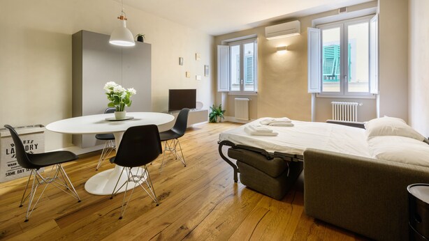 A modern hotel room with a bed, a dining table with chairs, a small plant, and a window with blinds. - Dimora Santa Croce by Mmega (Florence)