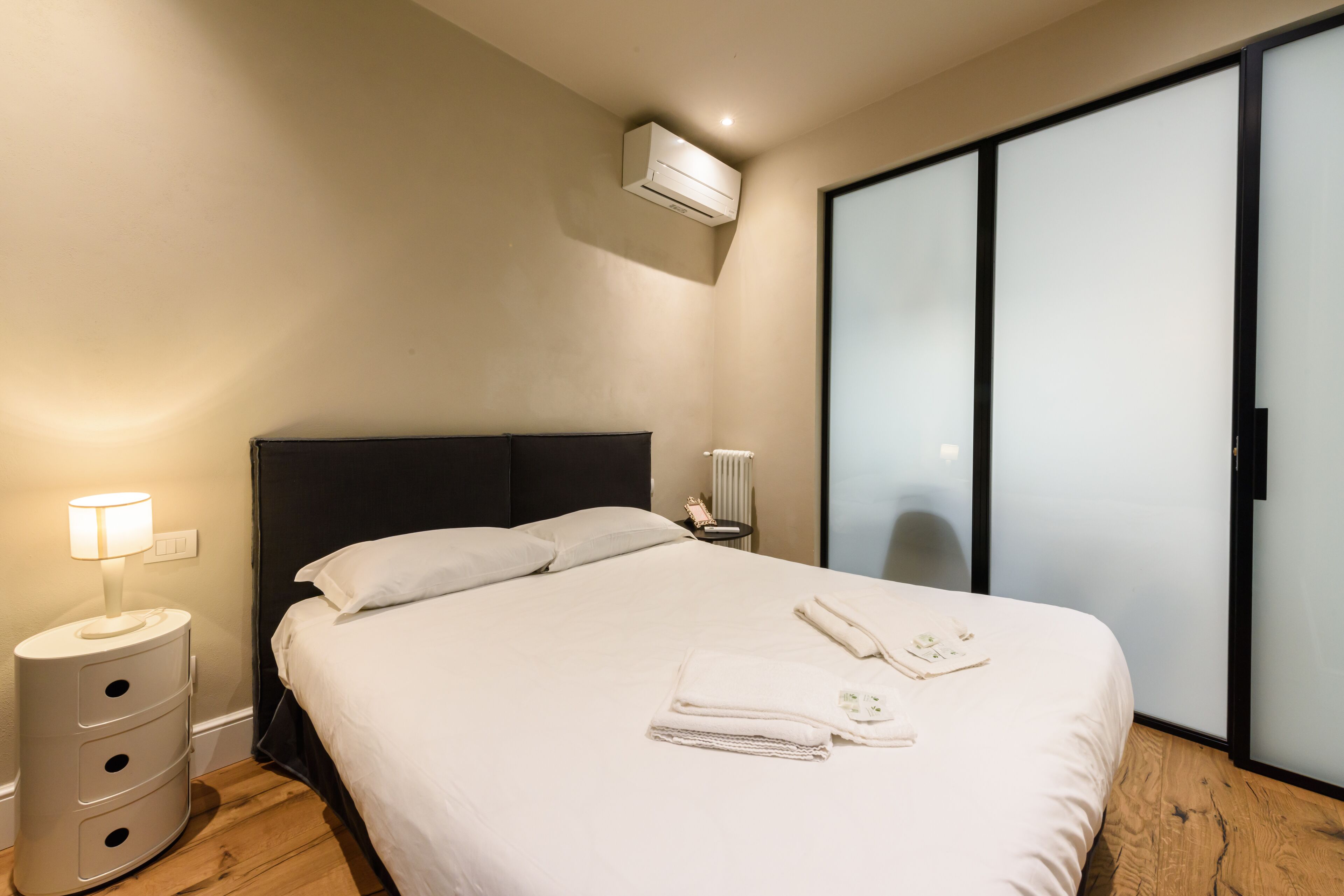 A modern hotel room with a bed, bedside lamp, air conditioning unit, and a wardrobe.