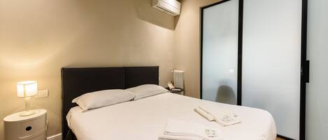 A modern hotel room with a bed, bedside lamp, air conditioning unit, and a wardrobe.