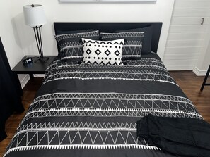 Ein Bett mit einer schwarz-weißen geometrischen Bettdecke, einer schwarzen Tagesdecke und einem Nachttisch mit Lampe und digitaler Uhr.