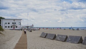 A beach with a wooden walkway leading to a white building with a lifeguard tower. - Bob W Pärnu (Parnu)