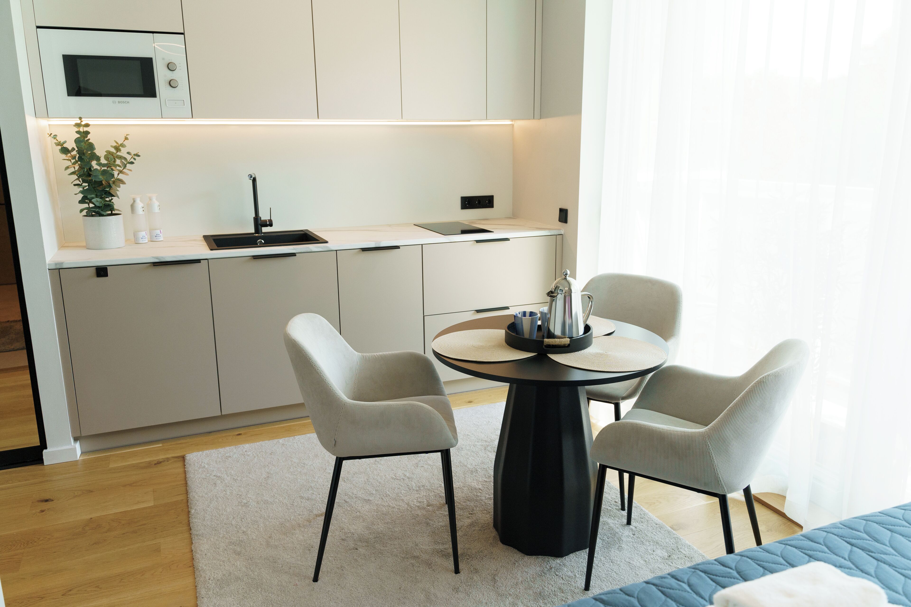 Una cocina moderna con microondas, lavamanos y encimera. Hay una zona de comedor con mesa y sillas, y una planta en maceta sobre la encimera.