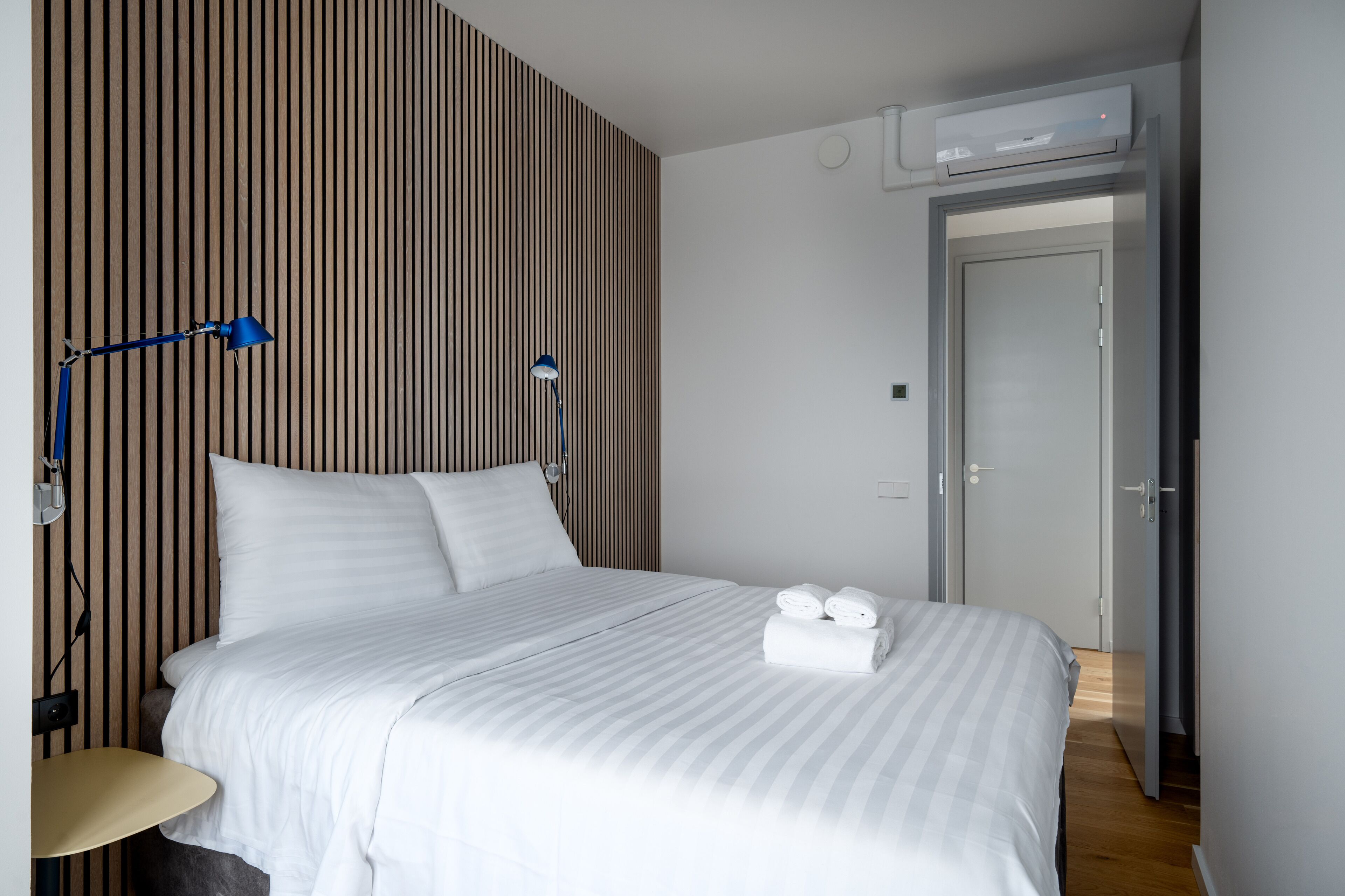 A hotel room with a bed, a desk, a lamp, and an air conditioning unit.