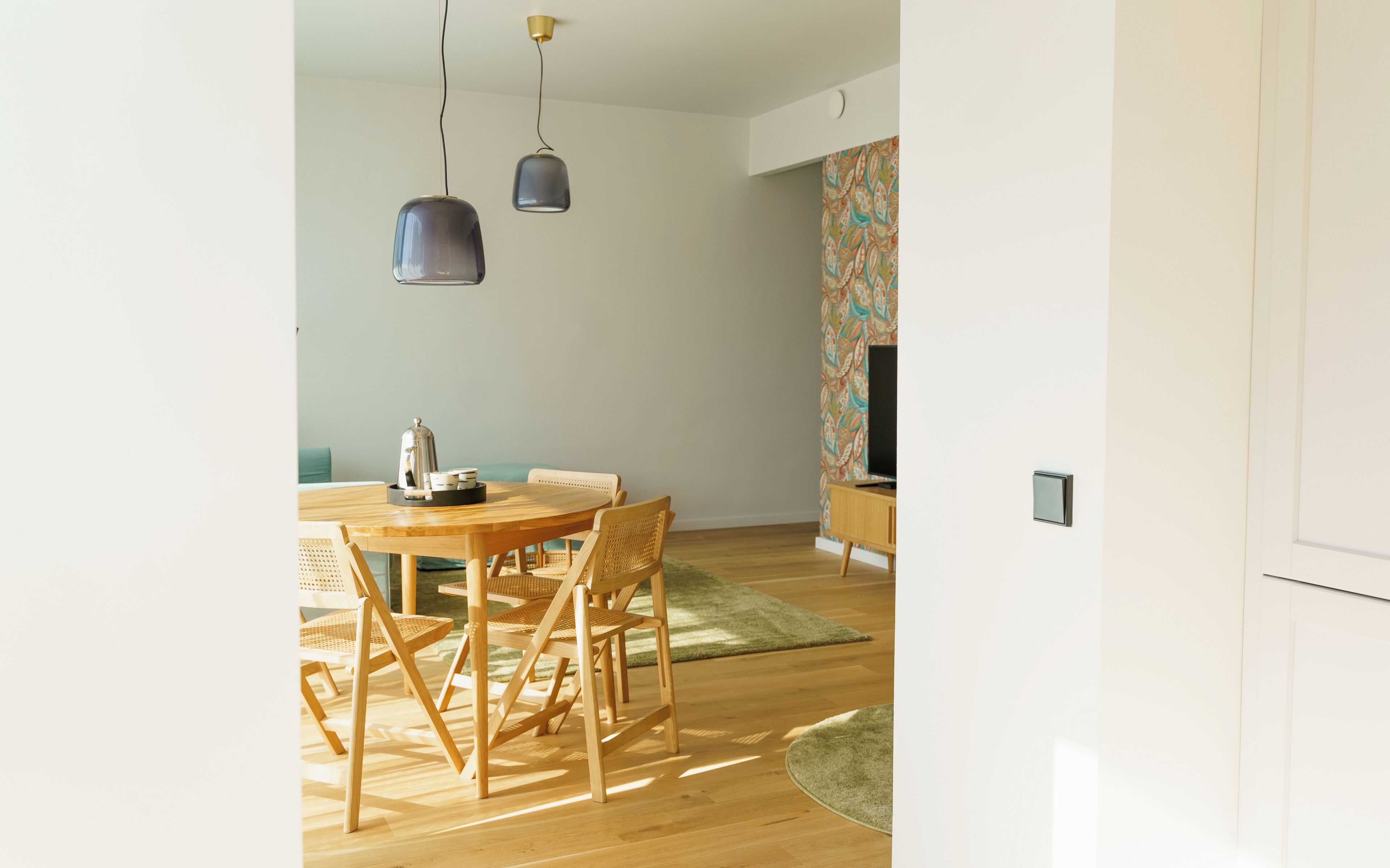 Un espace de salle à manger moderne, avec une table et des chaises en bois, des luminaires suspendus et un mur orné de motifs.