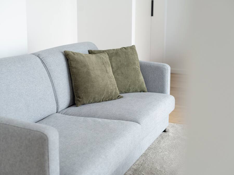 Un sofá gris con dos almohadas verdes en una habitación con paredes blancas y suelo de madera.