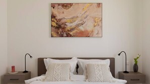 Une chambre à coucher avec un grand lit, deux tables de chevet, un tableau abstrait accroché au mur et un vase avec une plante sur l’une des tables de chevet.