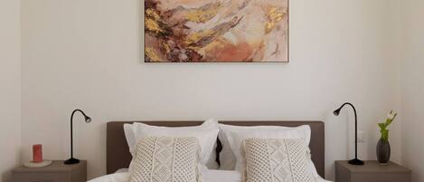 A bedroom with a large bed, two nightstands, a wall-mounted abstract painting, and a vase with a plant on one nightstand.
