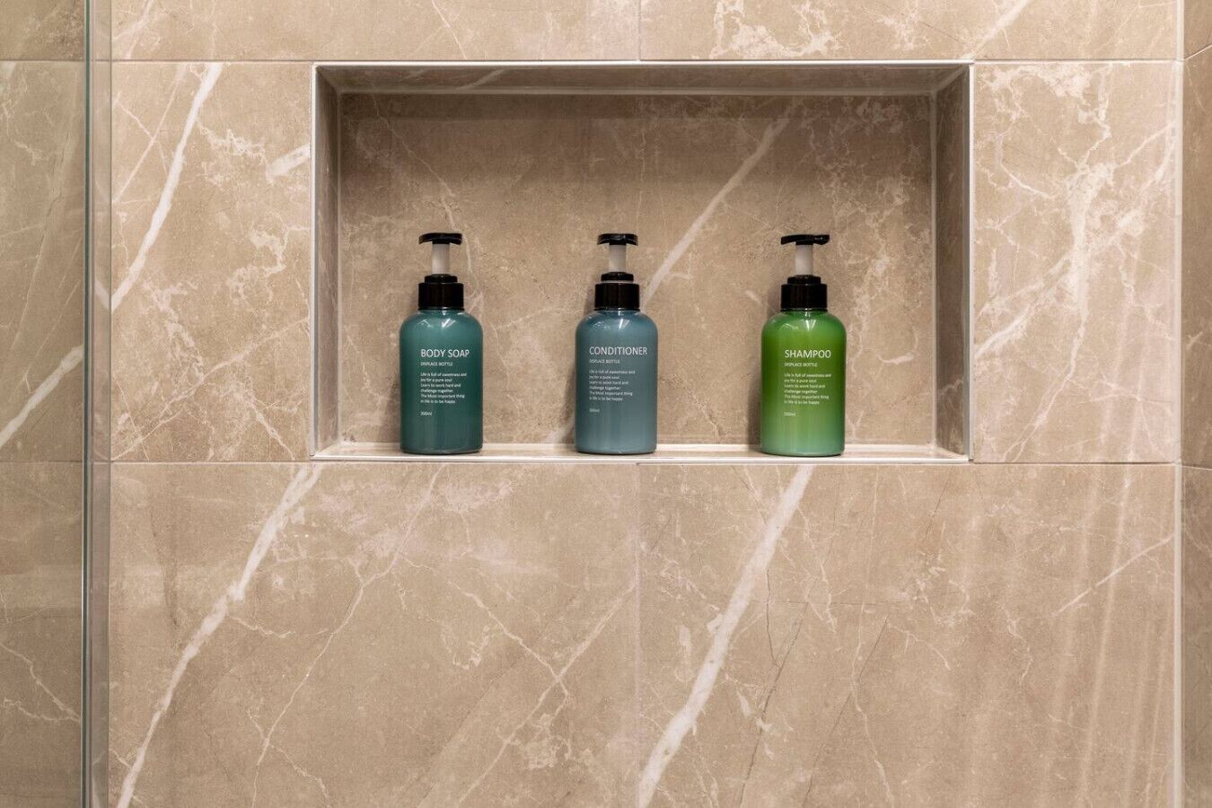 A bathroom with a marble wall and a shelf containing three bottles of toiletries: body soap, conditioner, and shampoo.