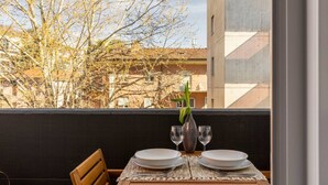A balcony with a table set for two, a vase with a single flower, and a view of buildings and trees.