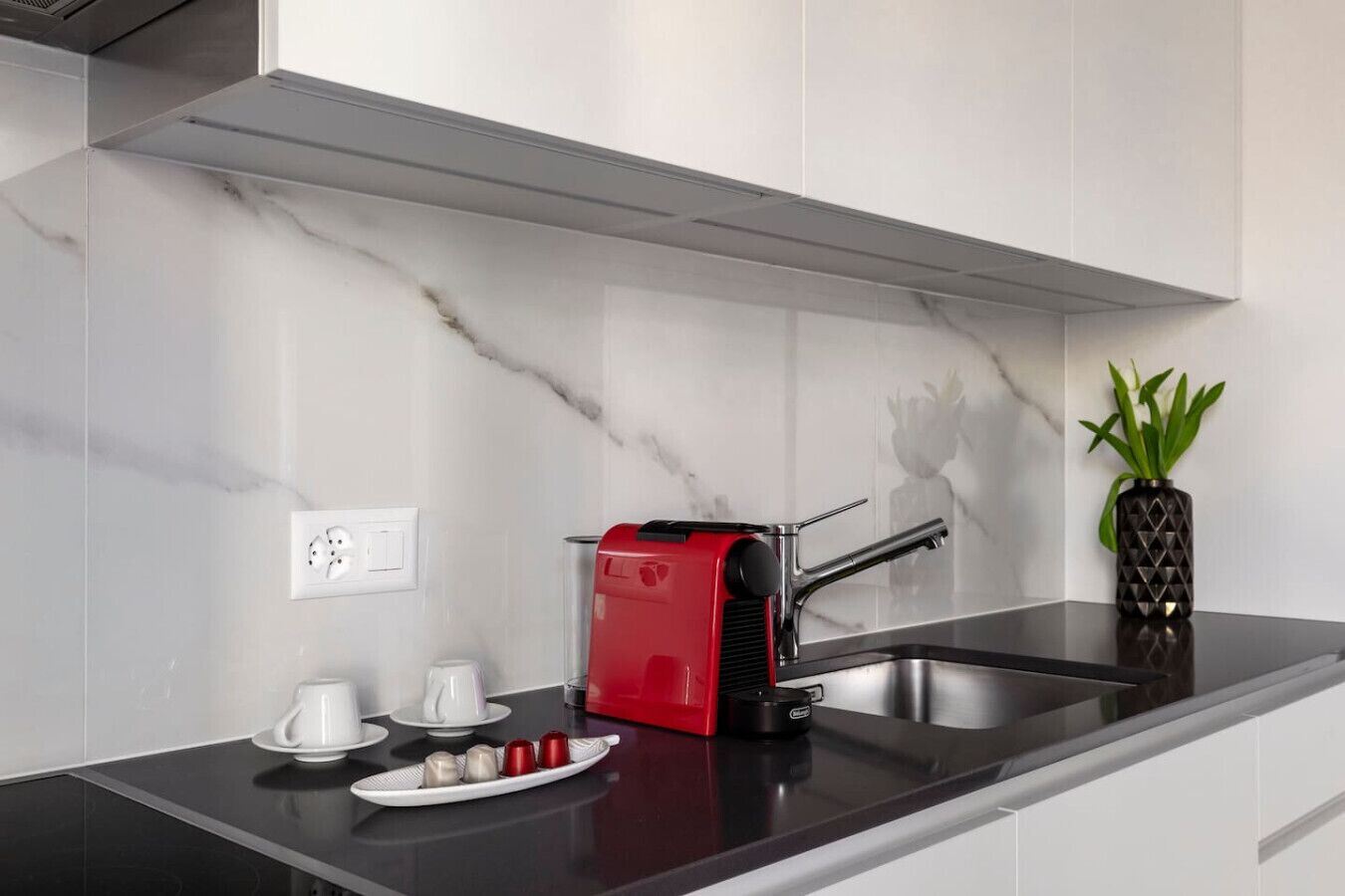 A modern kitchen with a black countertop, a red coffee machine, a stainless steel sink, and a potted plant.