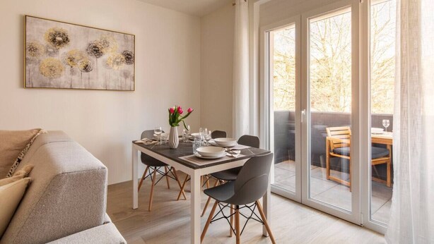 A dining area with a table set for two, a sofa, and a sliding glass door leading to an outdoor seating area.