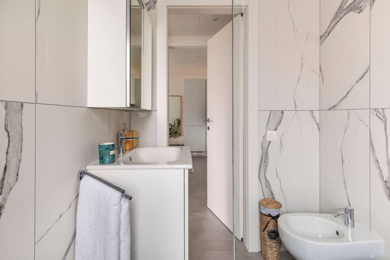 Un baño moderno con tocador blanco, lavamanos y bidé. Las paredes tienen un diseño de mármol y hay una toalla colgada en un perchero.