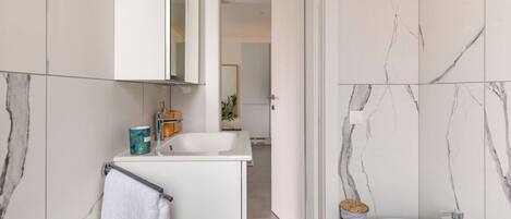 Un baño moderno con tocador blanco, lavamanos y bidé. Las paredes tienen un diseño de mármol y hay una toalla colgada en un perchero.