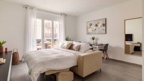 Une chambre à coucher moderne avec un lit, une table de chevet et un bureau avec une chaise. Il y a une grande fenêtre avec des rideaux, un tableau au mur et un miroir.