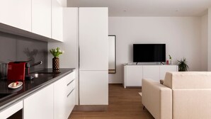 Une cuisine moderne avec des meubles blancs, un plan de travail noir et un appareil électroménager rouge. On y trouve un coin repas avec un canapé beige et un téléviseur à écran plat posé sur un meuble blanc.