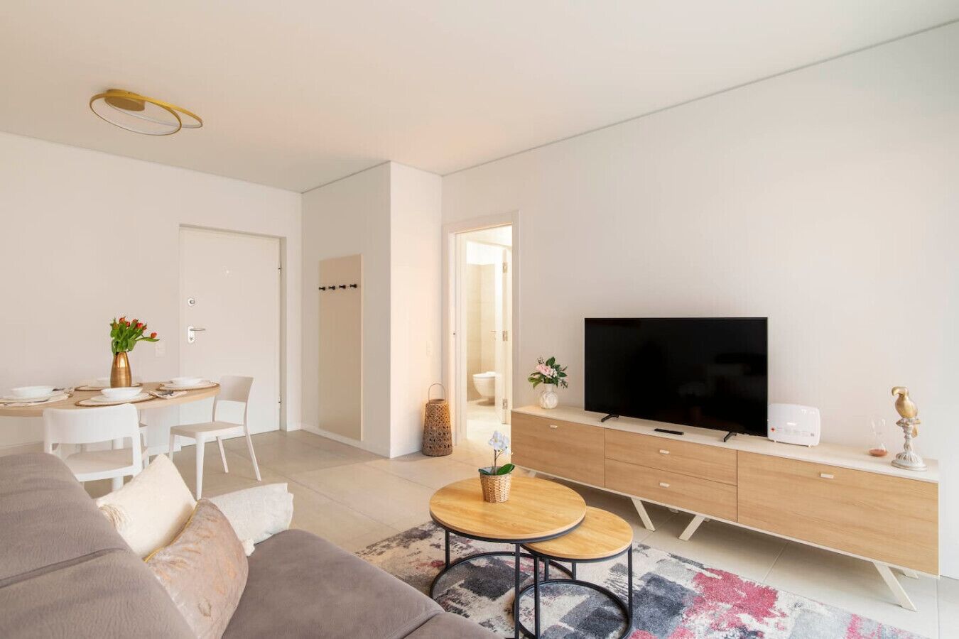 Ein modernes Wohnzimmer mit einer Couch, einem Esstisch mit Stühlen, einem Fernseher auf einer Holzkommode und einem kleinen runden Tisch mit einer Vase.