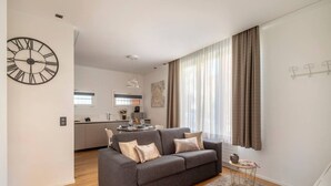 A modern living room with a grey sofa, a round wall clock, and a kitchen in the background. - Blue Diamond - Gorgeous lakefront apartment with parking (Lugano)