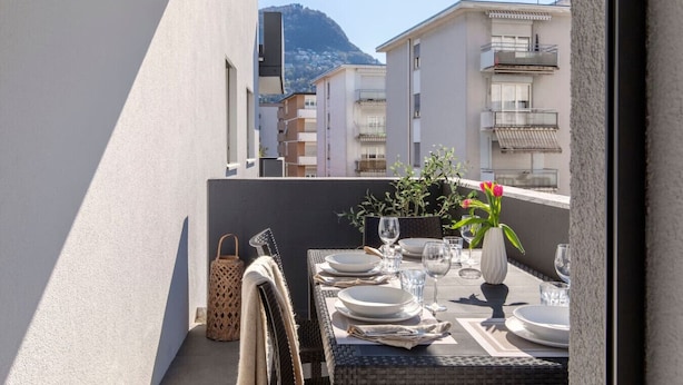 Un balcon avec une table dressée pour le dîner, des plantes en pot et une vue sur des immeubles et une montagne.