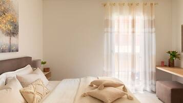 A bedroom with a large bed, a bedside table, a nightstand, a window with sheer curtains, and a painting on the wall.