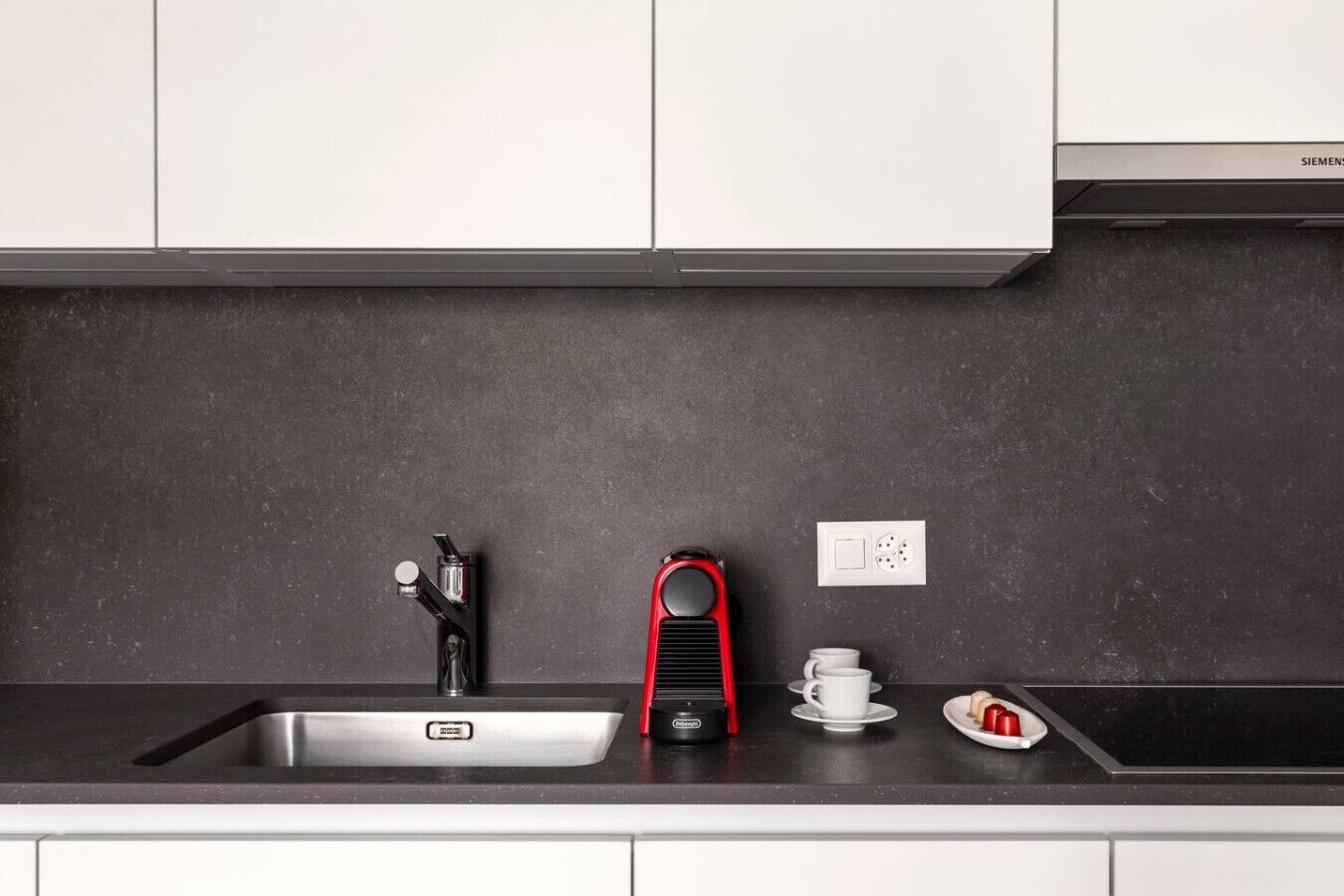 Uma cozinha moderna com uma pia de aço inoxidável, uma cafeteira vermelha, uma chávena branca e um prato com um pastel.