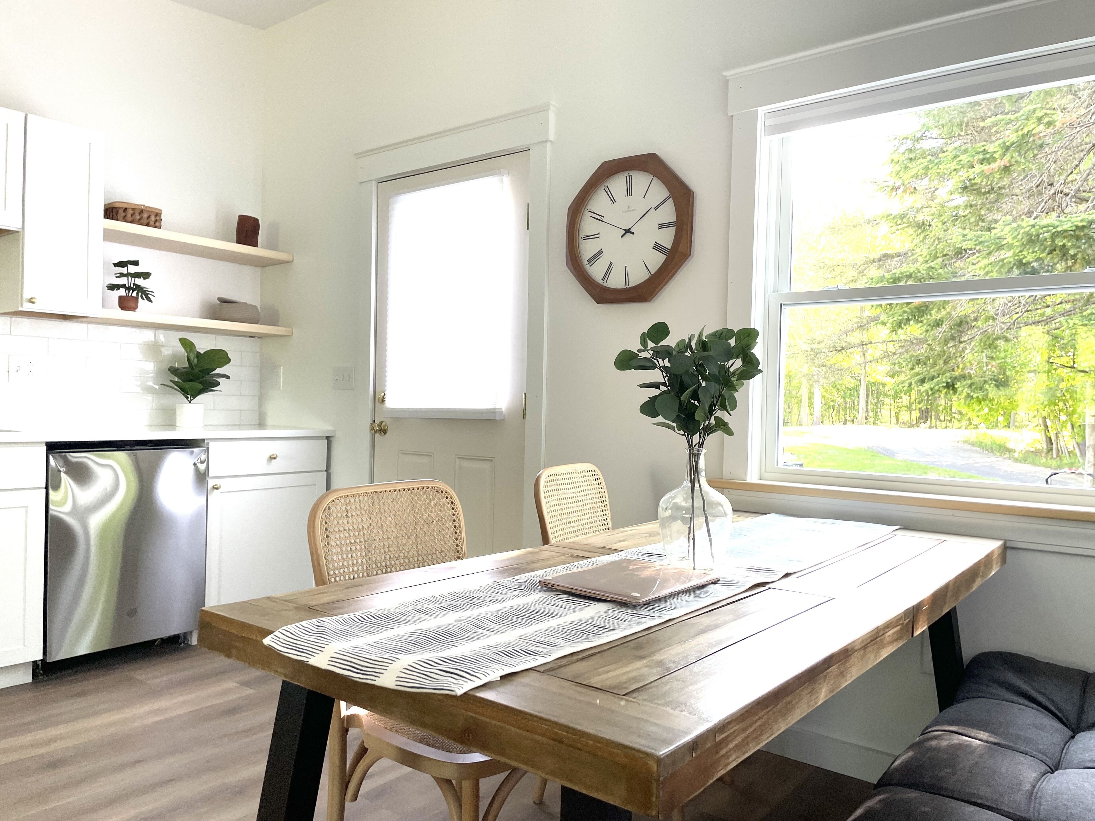 A kitchen with white cabinets, a stainless steel dishwasher, a wooden dining table with a placemat, wicker chairs, and a vase with green plants.