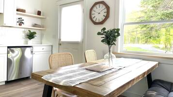 A kitchen with white cabinets, a stainless steel dishwasher, a wooden dining table with a placemat, wicker chairs, and a vase with green plants.