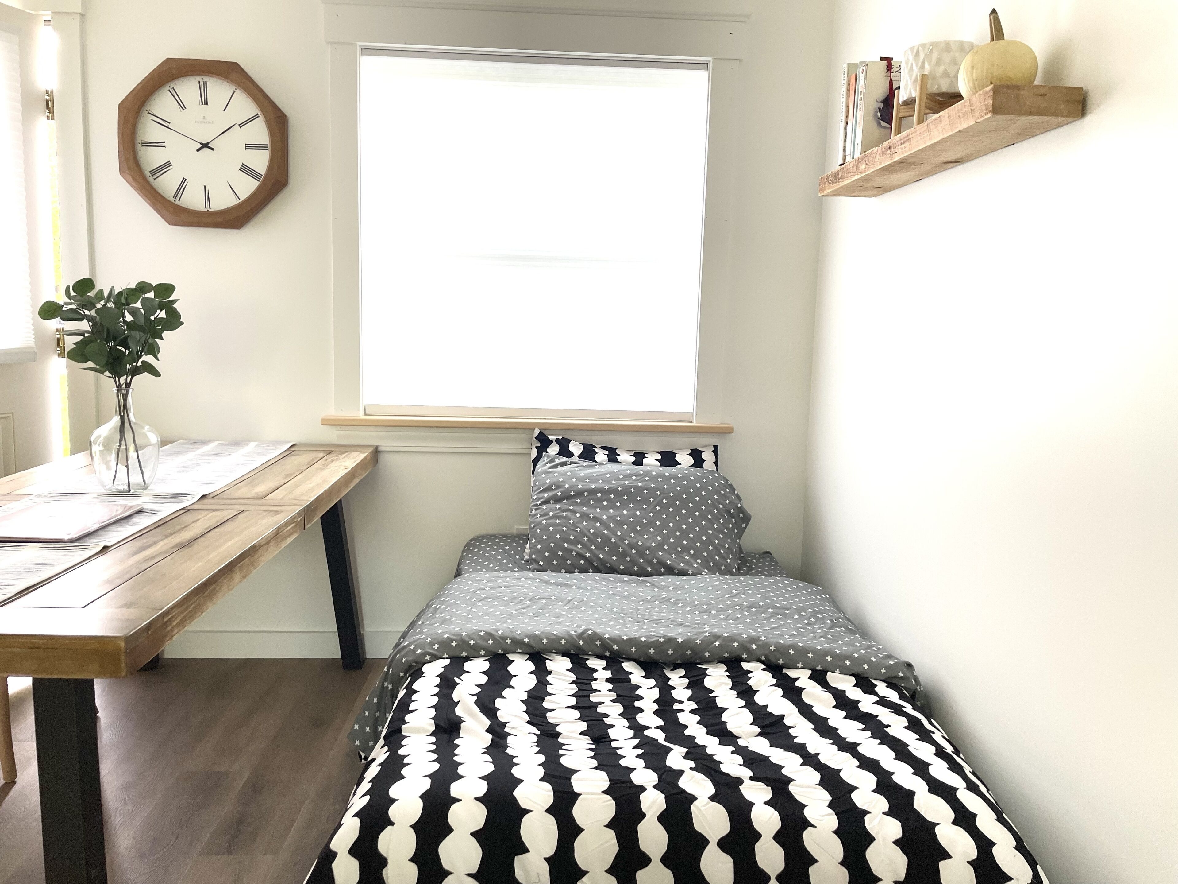 Un dormitorio con una cama de madera, un reloj de pared, una ventana y un pequeño estante con libros y una calabaza.