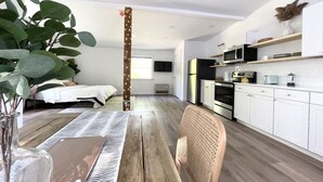 A modern kitchen with white cabinets, a microwave, and a stove. There is a dining table with a chair and a plant. - King Size Sun Rise Midcoast Lovely Stay (Topsham)