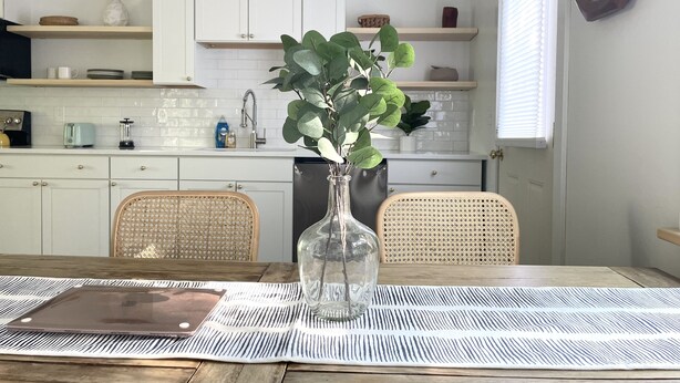 A modern kitchen with white cabinets, a wooden dining table, and a vase with green leaves. - King Size Sun Rise Midcoast Lovely Stay (Topsham)