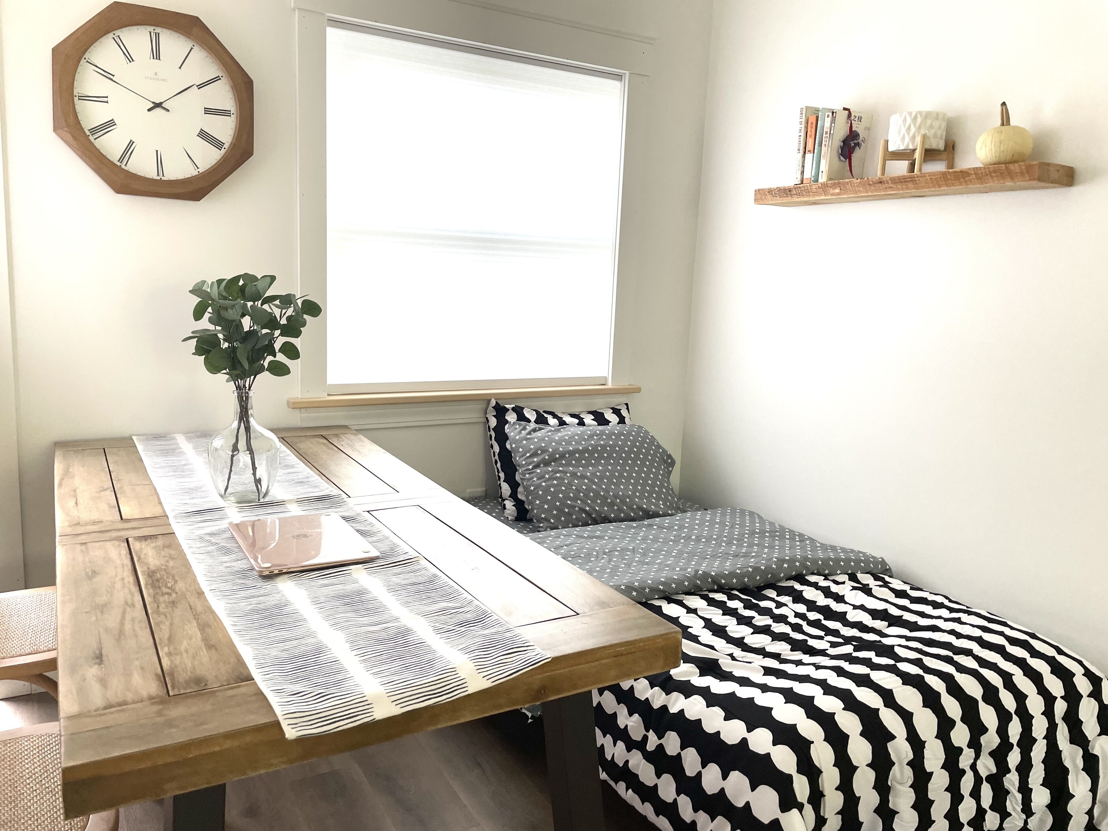 Quarto com cama de madeira, mesa de cabeceira com um vaso, relógio e janela com persianas.