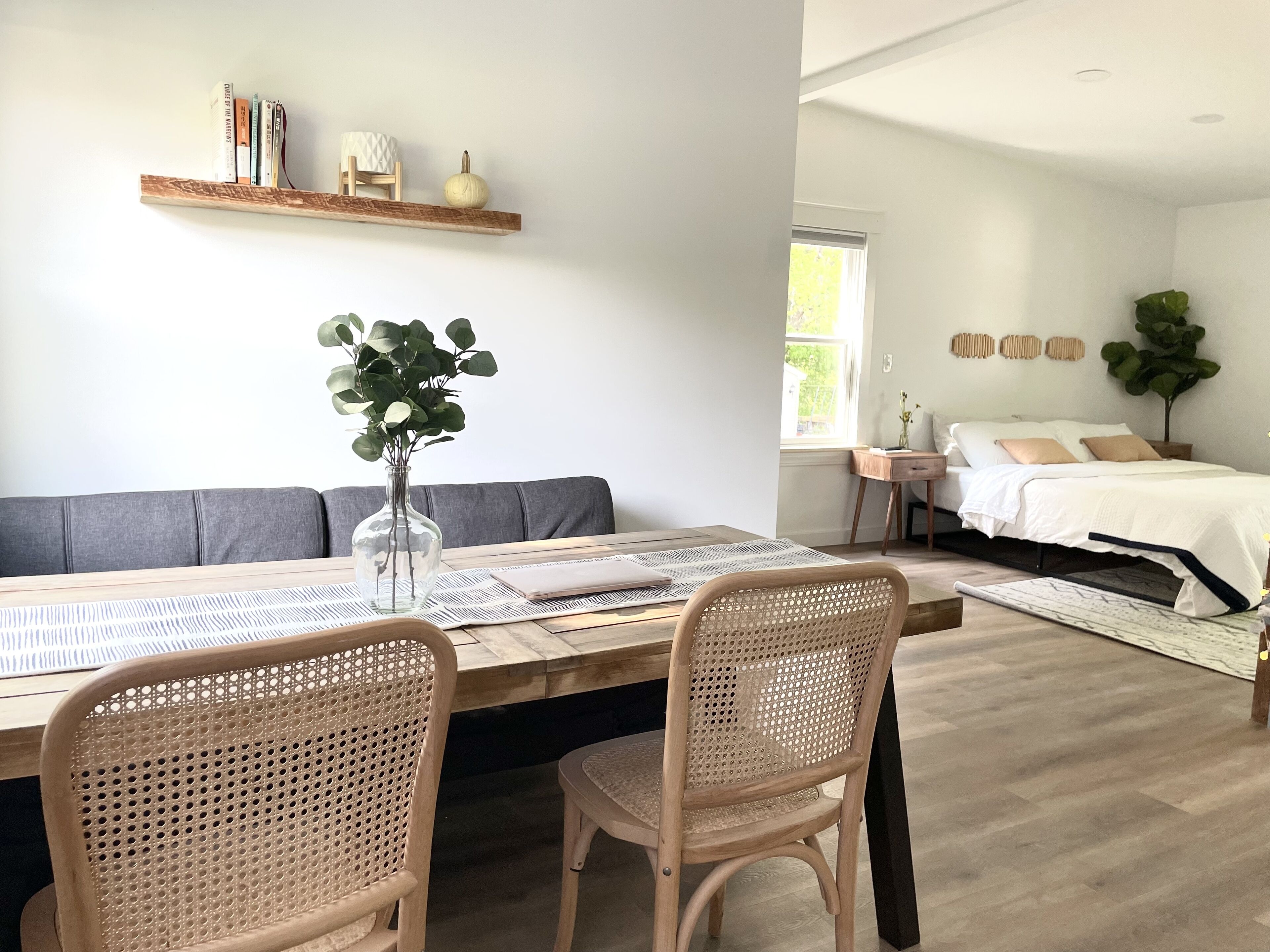 Una sala moderna con una mesa de comedor, dos sillas de mimbre, un sofá, una estantería y una cama con una mesita de noche.