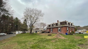 A two-story brick house with a large lawn, a tree, and a driveway with a parked car. - King Size Sun Rise Midcoast Lovely Stay (Topsham)