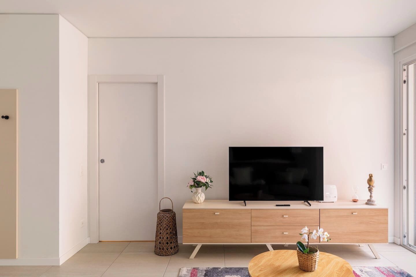Ett modernt vardagsrum med en platt-TV placerad på ett träskåp, ett litet bord med en vas med blommor och ett dekorativt ljus.