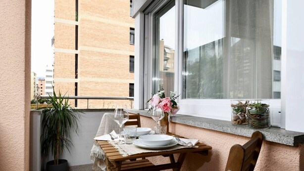 Un balcone con un tavolo di legno apparecchiato per due persone, una pianta in vaso e una finestra che si affaccia su edifici.