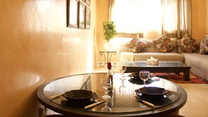 A dining area with a round glass table set for two, a sofa with floral cushions, and a window with curtains in the background. - Amoud apartments & flats (Marrakech)