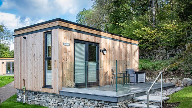 A modern wooden cabin with a stone foundation, a glass door, and a small outdoor seating area. - Rosthwaite  Woodland pod - a holiday Pod that sleeps 2 with an en-suite (Winster)