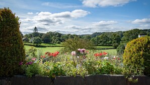 A garden with a stone wall, various flowers, and a view of a green landscape with trees and a house in the background. - Rosthwaite  Woodland pod - a holiday Pod that sleeps 2 with an en-suite (Winster)