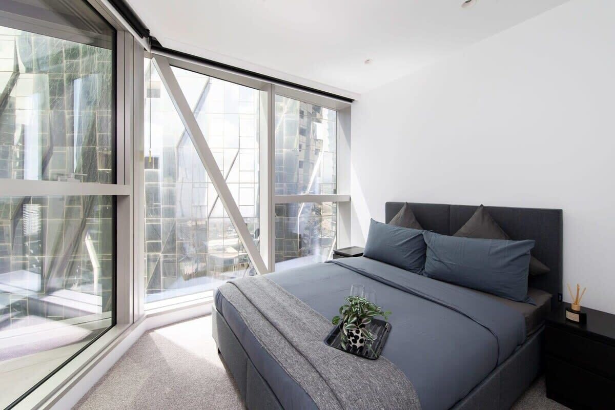 Een moderne slaapkamer met een groot raam, een bed met grijze beddengoed en een klein plantje op het bed.