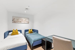 Une chambre avec deux lits, chacun doté d’une tête de lit bleue et de draps blancs, un petit bureau et une chaise.