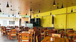 A spacious dining area with wooden tables and chairs, a high ceiling with hanging lights, and a yellow wall with signage. - Bay Breeze Hotels & Resorts (Morjim)