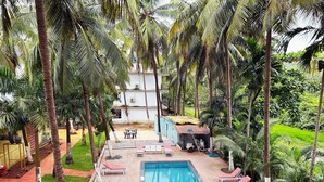 A swimming pool with lounge chairs, surrounded by palm trees and a building in the background. - Bay Breeze Hotels & Resorts (Morjim)