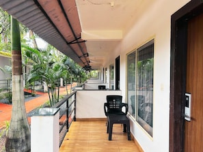 Un balcón cubierto con piso de madera, sillas de plástico negro y vista a un jardín con palmeras.
