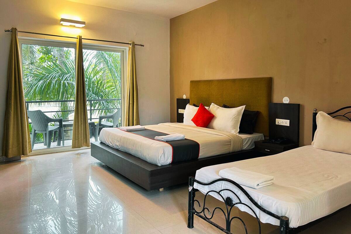 Habitación de hotel con una cama grande, otra más pequeña, un balcón con una silla y vistas al verdor exterior.