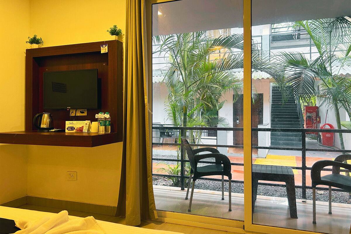 Habitación de hotel con cama, televisor montado en la pared, un pequeño estante con artículos de tocador y vista al área exterior con sillas y plantas.