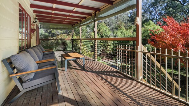 A covered outdoor seating area with a wooden bench and a table, surrounded by a wooden railing and a view of trees and foliage.